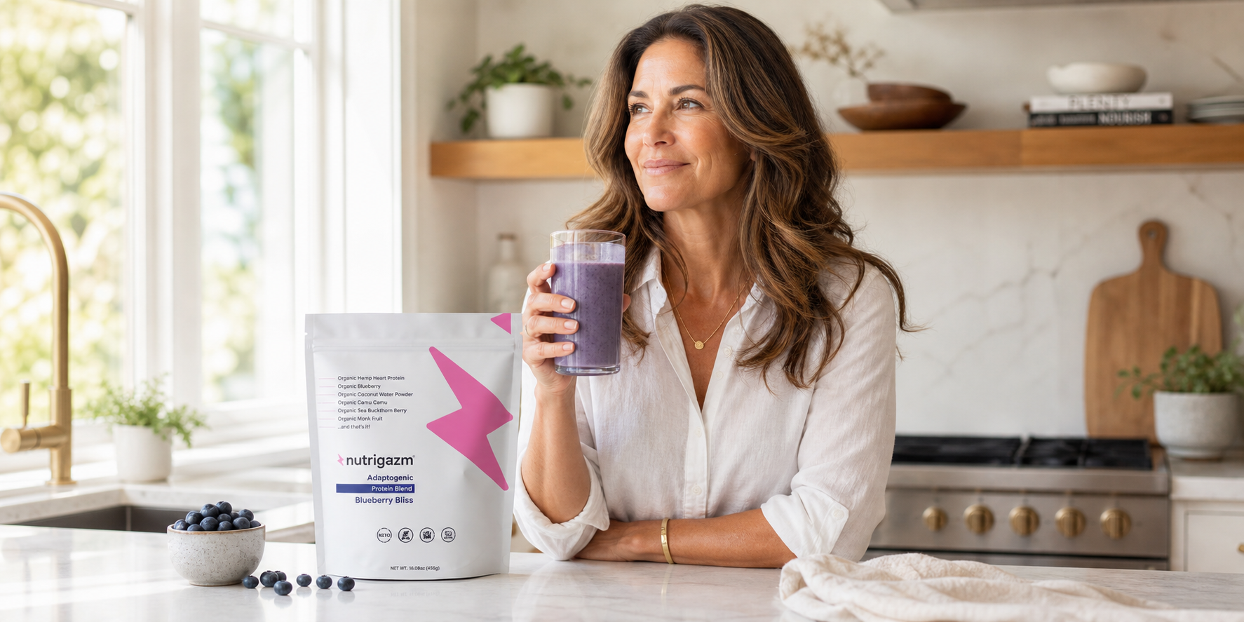 Woman in natural kitchen light holding a blueberry Nutrigazm smoothie beside the Blueberry Bliss pouch