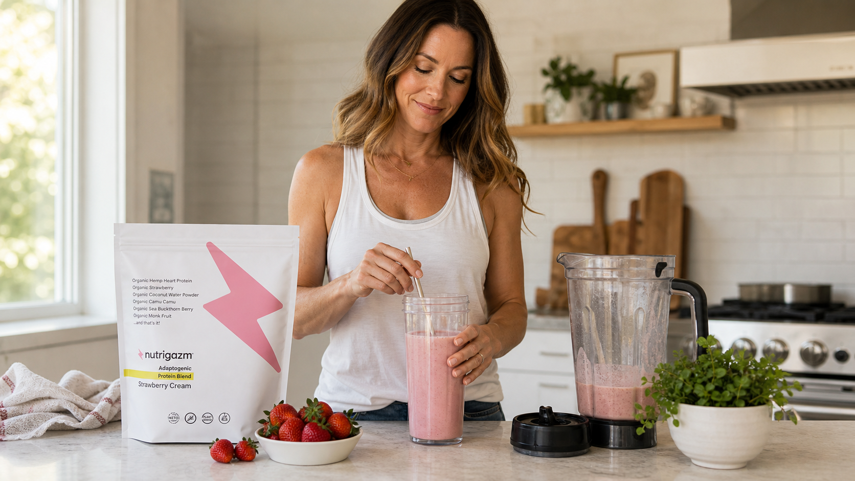 Woman in natural kitchen light stirring a creamy strawberry Nutrigazm smoothie beside the Strawberry Cream pouch