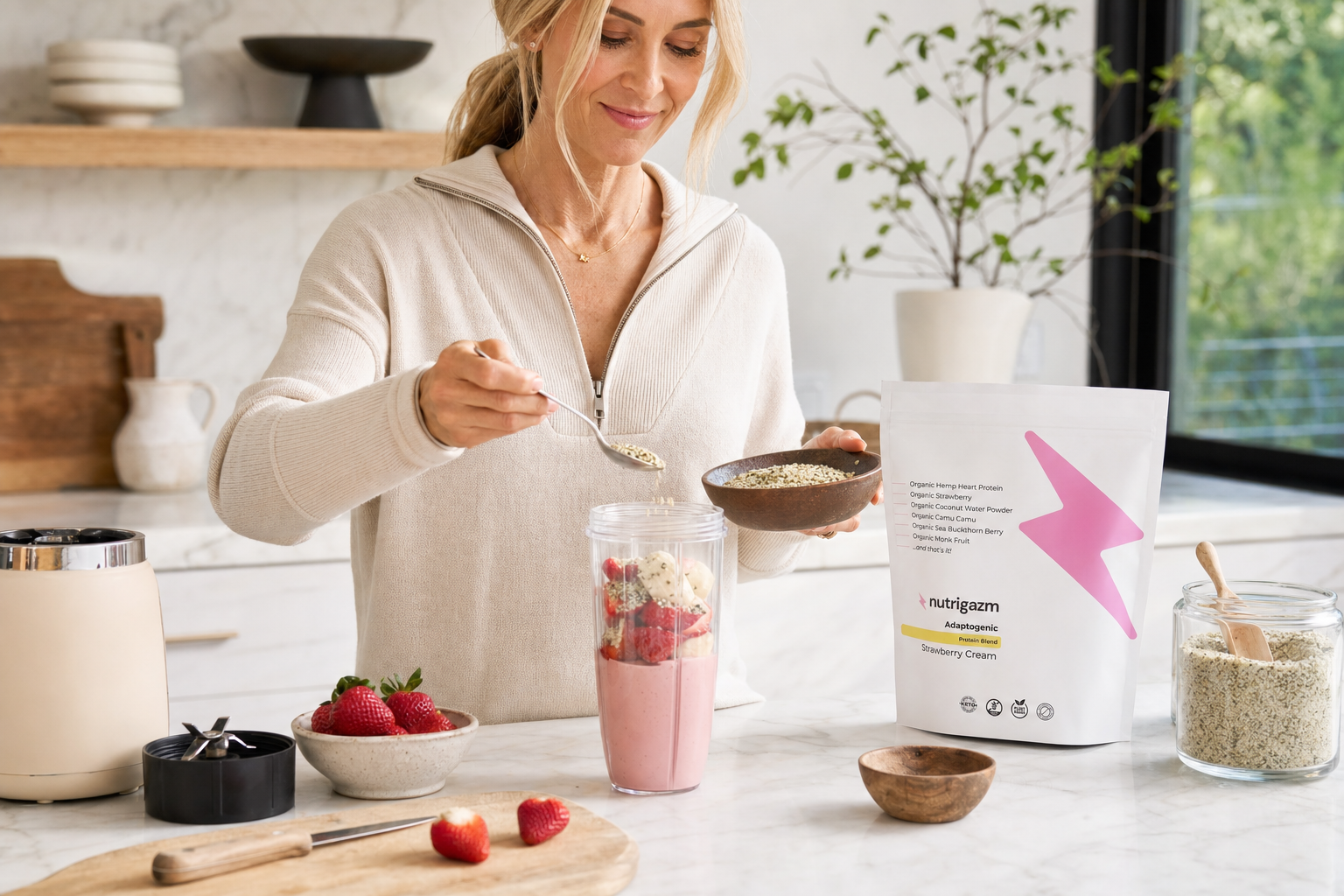 Woman in natural kitchen light adding hemp hearts to a strawberry Nutrigazm smoothie beside the Strawberry Cream pouch